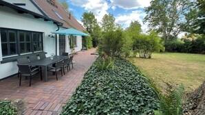 Une terrasse avec une table et des chaises, un bâtiment blanc avec un toit en tuiles et un jardin agrémenté d’une haie.