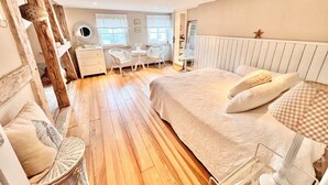 Une chambre avec un plancher en bois, un lit avec une literie blanche, une commode, une chaise et un bureau avec un miroir.