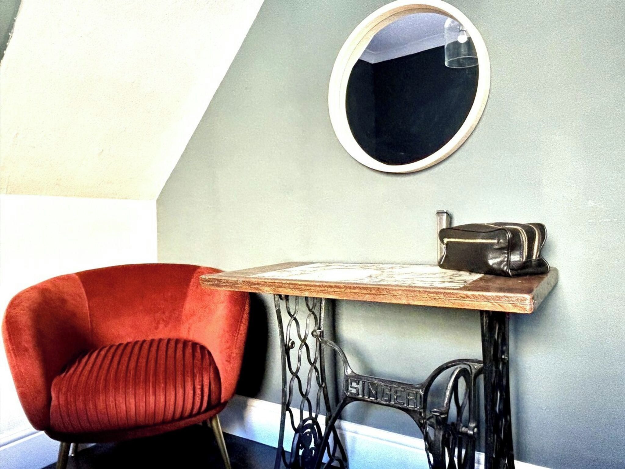 A room with a red chair, a sewing machine, and a round mirror on the wall.