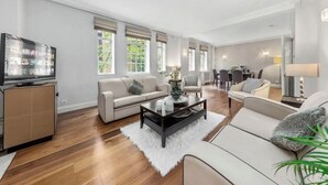 Ein modernes Wohnzimmer mit einer Couch, einem Couchtisch und einem Fernseher. Im Hintergrund sind große Fenster und ein Essbereich zu sehen.