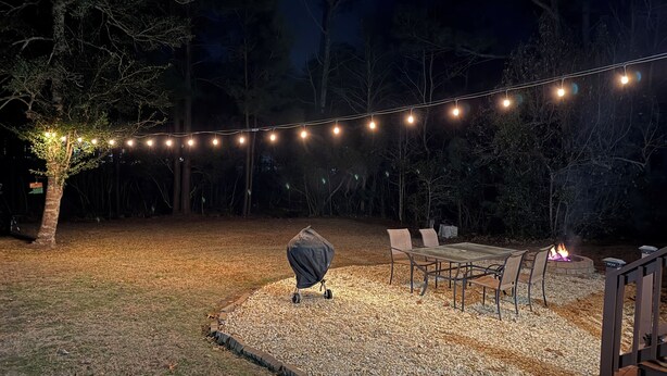 Un espace extérieur aménagé avec un foyer, des guirlandes lumineuses et un barbecue couvert.