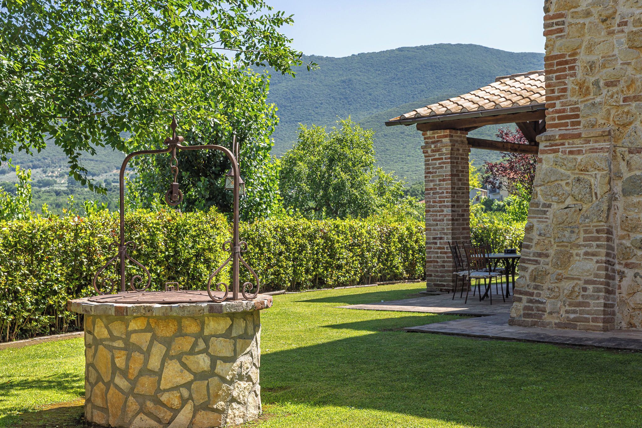 A stone well with a metal structure, a brick pergola, and a table with chairs in a garden with a mountain backdrop.