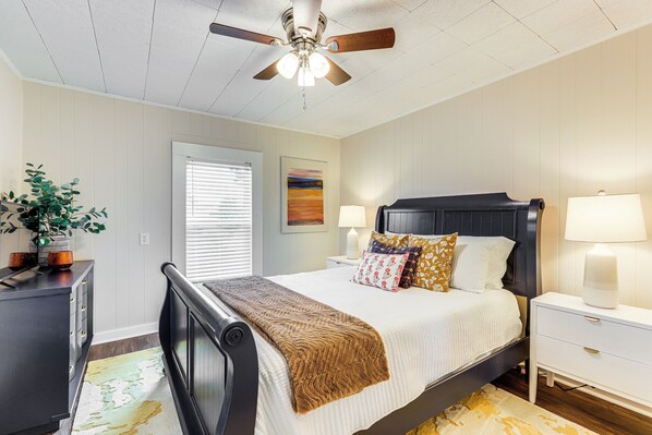 A bedroom with a bed, bedside tables, a ceiling fan, a window with blinds, and a painting on the wall.