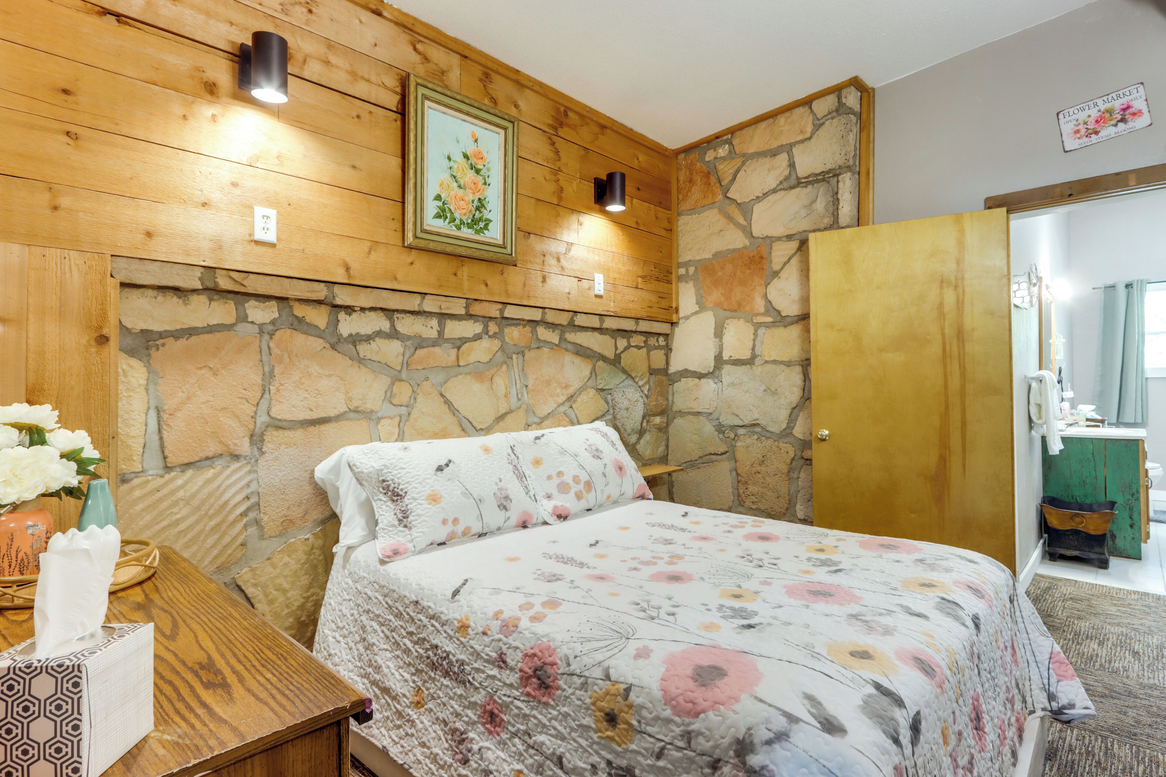 A bedroom with a stone wall, wooden ceiling, a floral bedspread, and a painting above the bed.