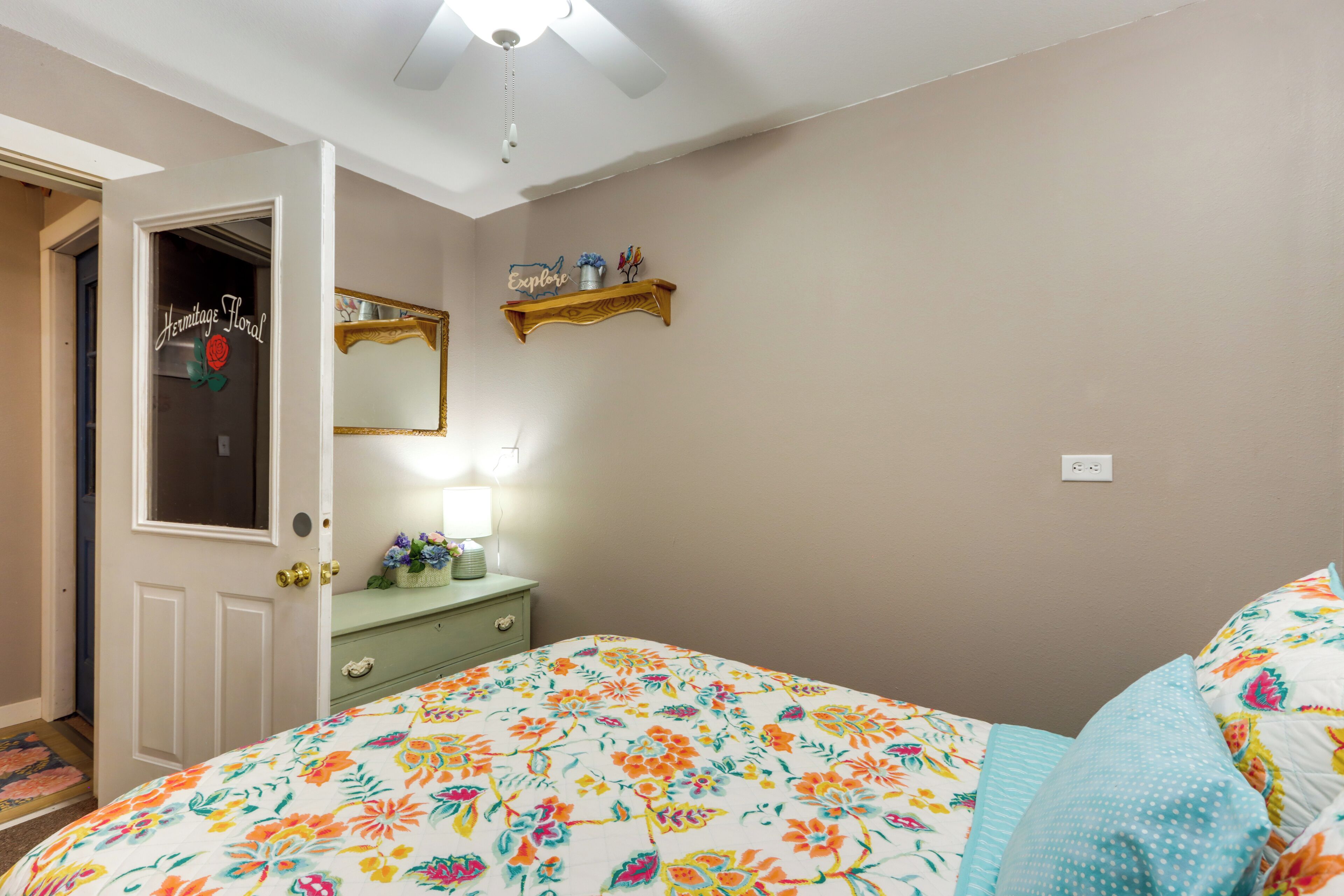 A bedroom with a floral bedspread, a dresser with a mirror, and a shelf with decorative items.