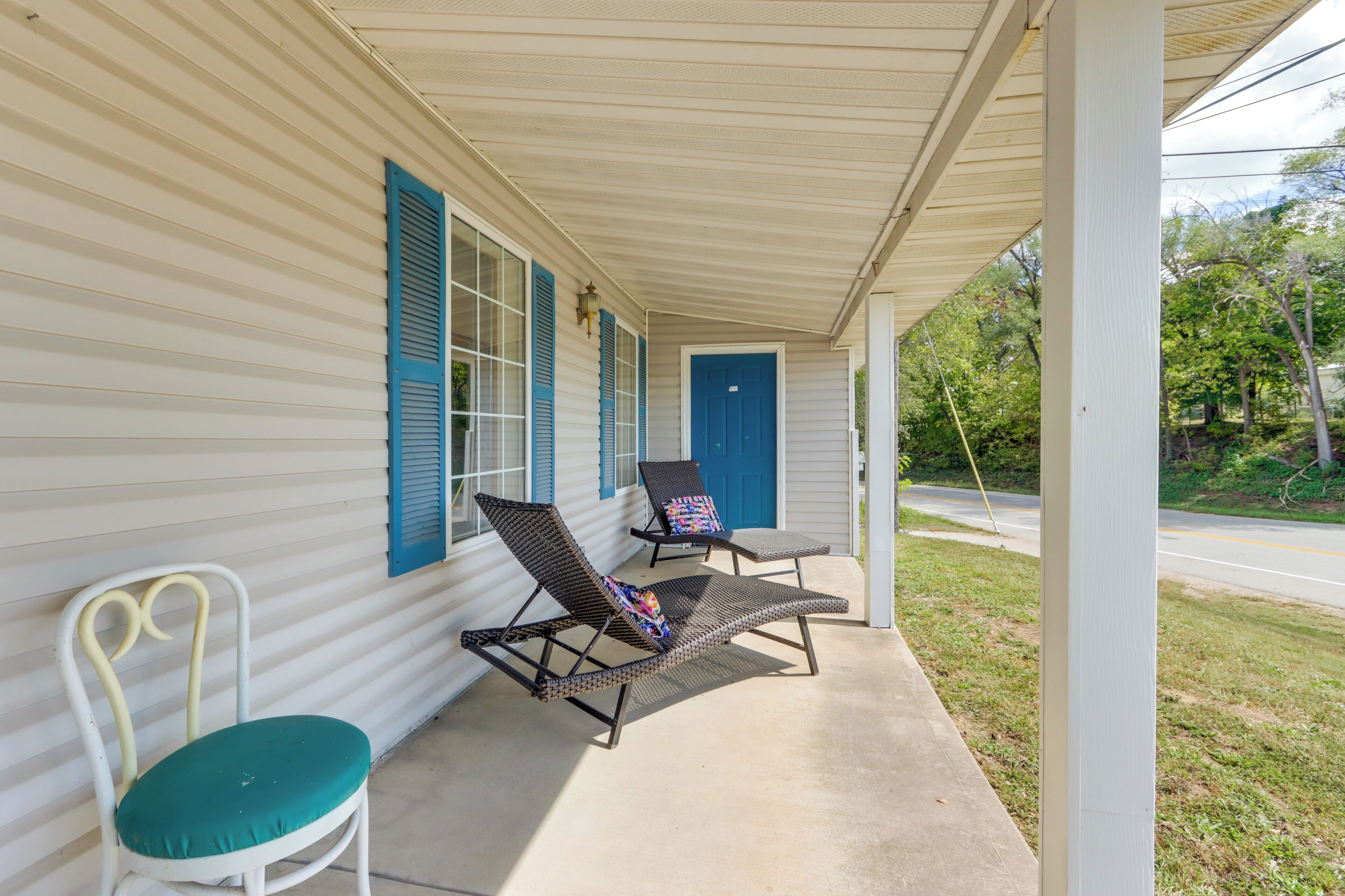 A covered porch with two lounge chairs and a white chair, a blue door, and a window with blue shutters.