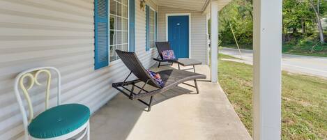 A covered porch with two lounge chairs and a white chair, a blue door, and a window with blue shutters.
