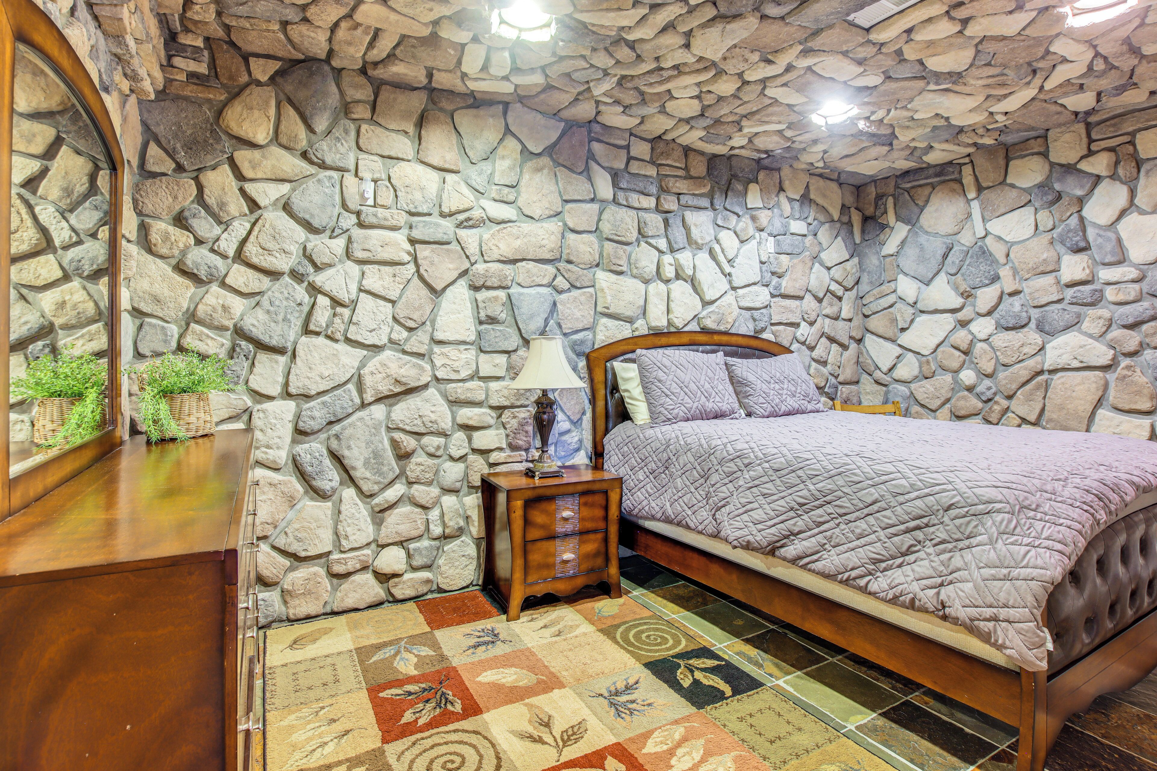 A bedroom with a stone wall, a bed with a quilt, a nightstand, and a mirror.