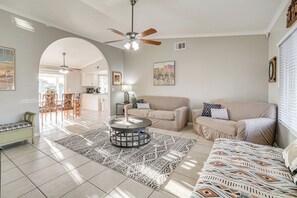 A spacious living room with a beige sofa, a round coffee table, and a patterned rug. There is a ceiling fan, a dining area in the background, and a kitchen visible through an archway. - 8 Mi to State Farm Stadium: Glendale Home w/ Grill (Peoria)