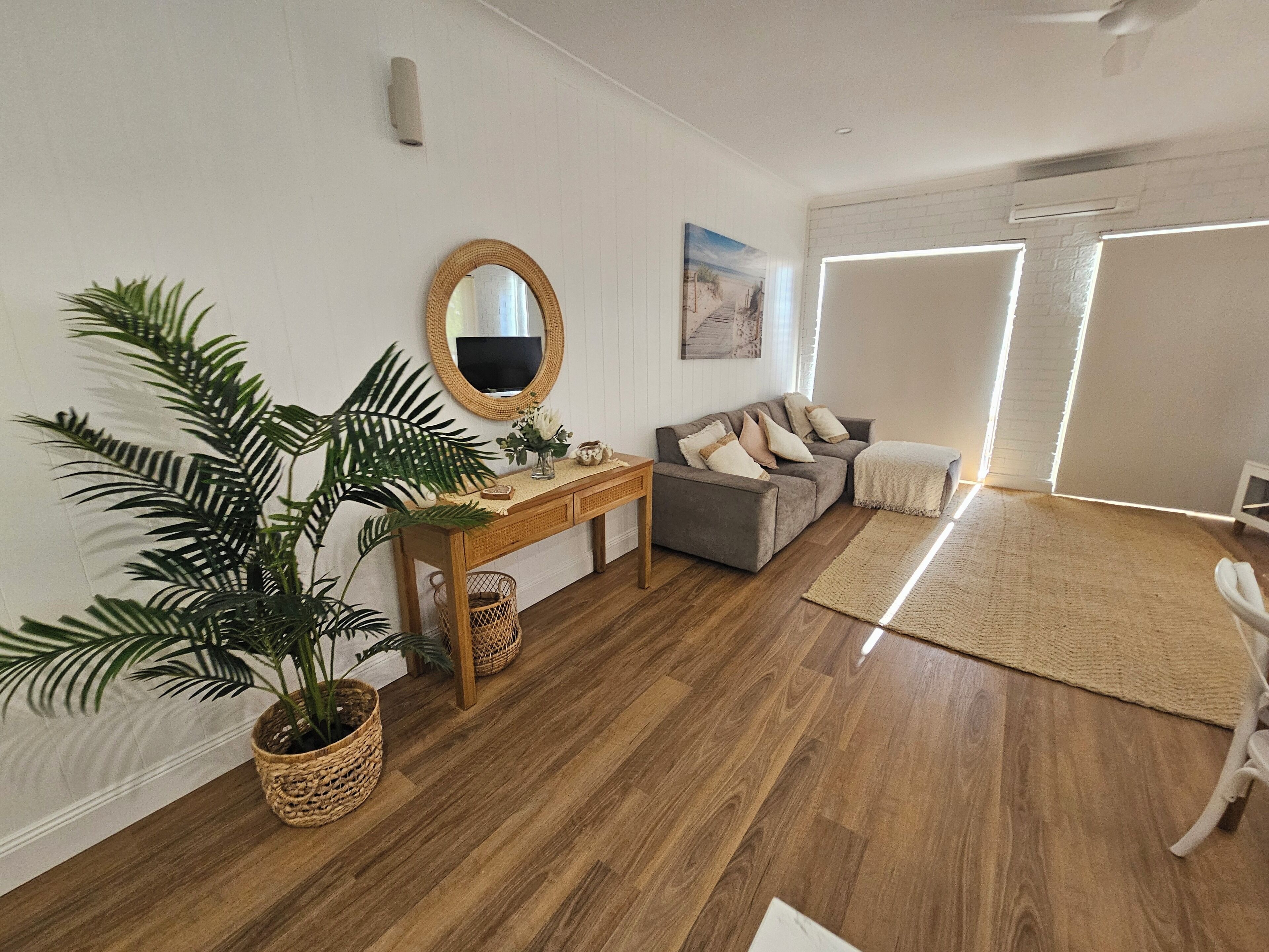 A modern living room with a wooden floor, a grey sofa, a wooden console table, a large potted plant, and a white armchair.