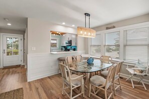 A dining area with a glass table and wicker chairs, a kitchen in the background, and a 'Beach' sign on the wall.