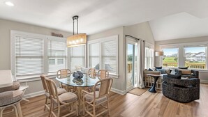 A spacious dining area with a glass table and wicker chairs, large windows with blinds, and a view of a residential area. - Exhale: Renovated, Hot Tub, Golf Course Views (Nags Head)