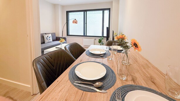 A dining area with a wooden table set for four, including plates, cutlery, and glasses. There is a window with a view of the outside, a sofa, and a lamp in the background. - London's 1-Bedroom Haven near The Shard in Elephant & Castle (London)