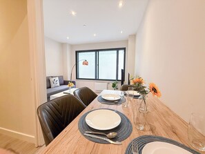 Comedor con mesa de madera dispuesta para cuatro, con platos, cubiertos y vasos. Se ve una ventana con vistas al exterior, un sofá y una lámpara al fondo.