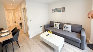 A modern living room with a grey sofa, white coffee table, and black chairs. There are framed artworks on the wall and a hallway leading to another room. - London's 1-Bedroom Haven near The Shard in Elephant & Castle (London)