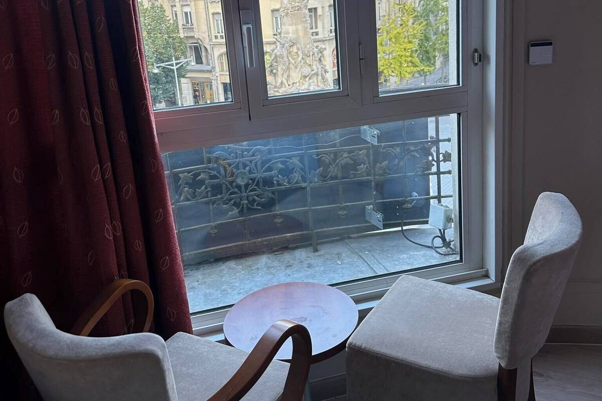 Une pièce dotée d’une grande fenêtre, de deux chaises, d’une petite table et offrant une vue sur les bâtiments environnants.