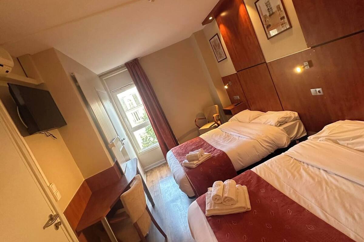 Une chambre d’hôtel avec deux lits, un bureau et une chaise. Il y a une fenêtre avec des rideaux, un téléviseur à écran plat fixé au mur et une porte qui donne sur une autre pièce.