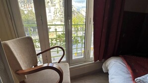 Ein Zimmer mit einem Holzsessel, einem Bett mit roter Decke und einem großen Fenster mit Blick auf ein Gebäude mit Statuen.