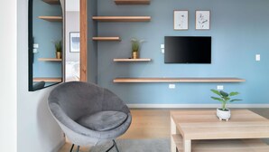 A modern living room with a grey armchair, a wooden coffee table, a flat-screen TV mounted on the wall, and several wooden shelves with plants. - w Spectacular Loft w Balcony in El Nogal (Bogotá)