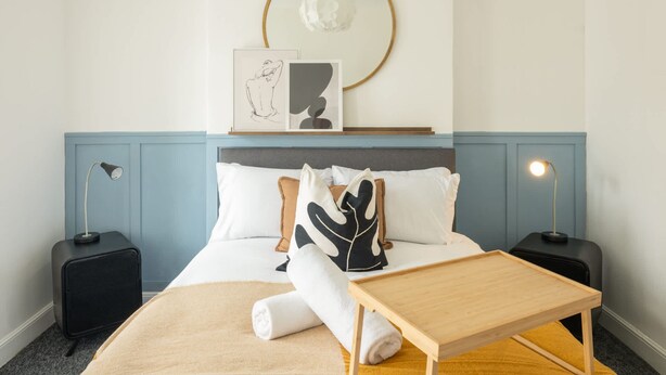 A neatly arranged hotel room with a bed, bedside tables, a bedside lamp, a small wooden table, and a wall-mounted artwork. - Host Stay Craddock Street (Cardiff)