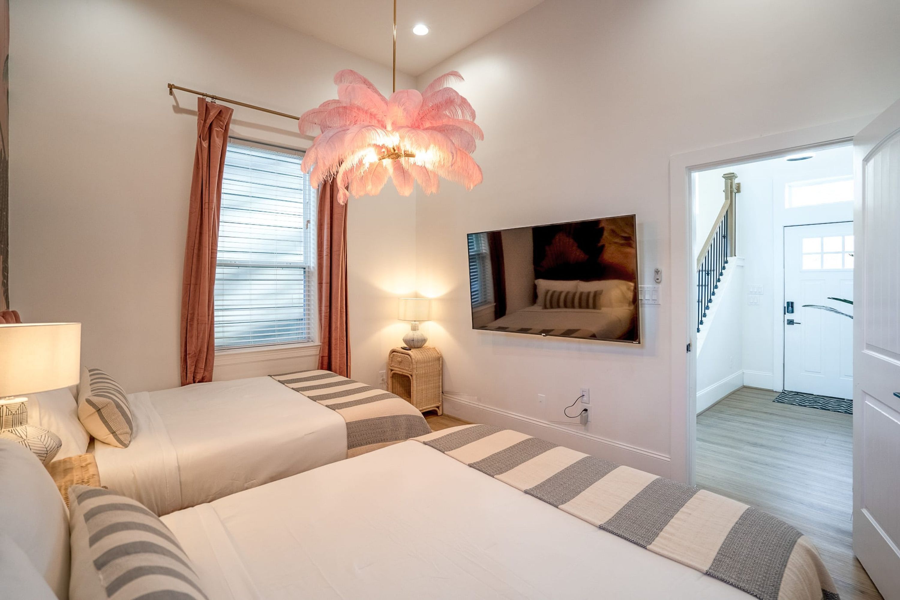 A bedroom with two beds, a large feathered chandelier, a painting on the wall, and a staircase leading to the door.