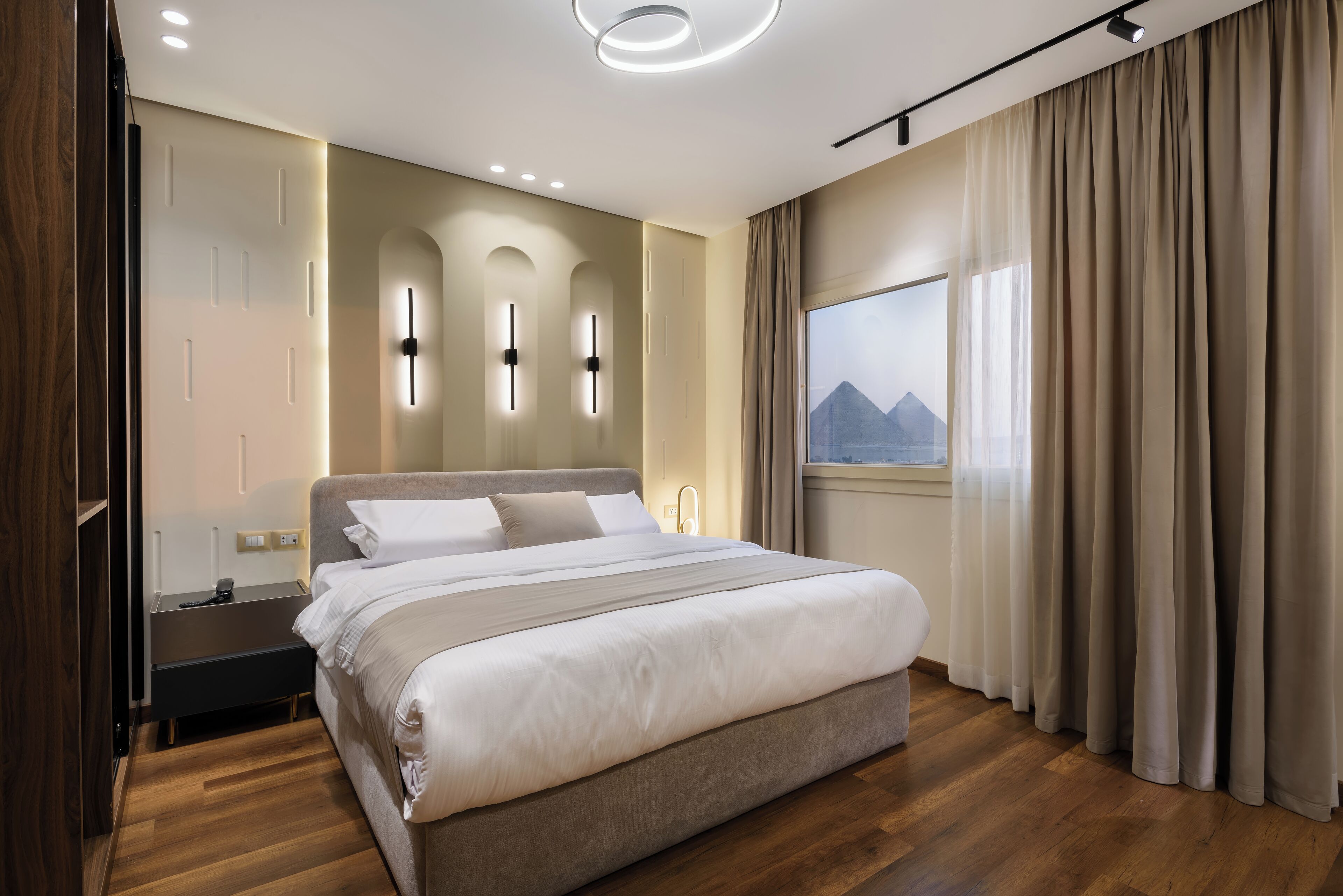 A modern hotel room with a large bed, a nightstand, a window with curtains, and a view of mountains.