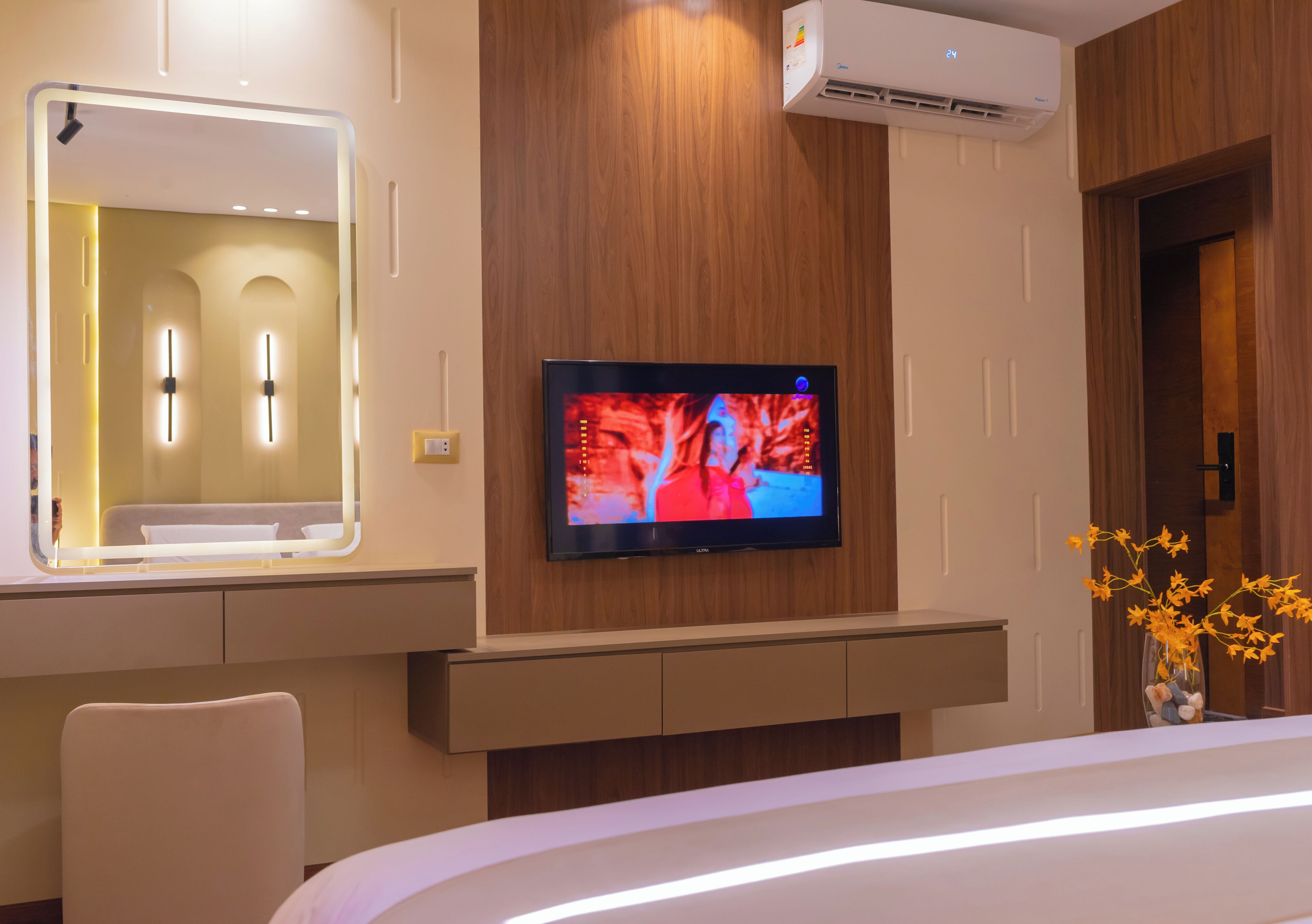 A modern hotel room with a flat-screen TV mounted on the wall, a large mirror above a vanity, and a vase with yellow flowers on a side table.