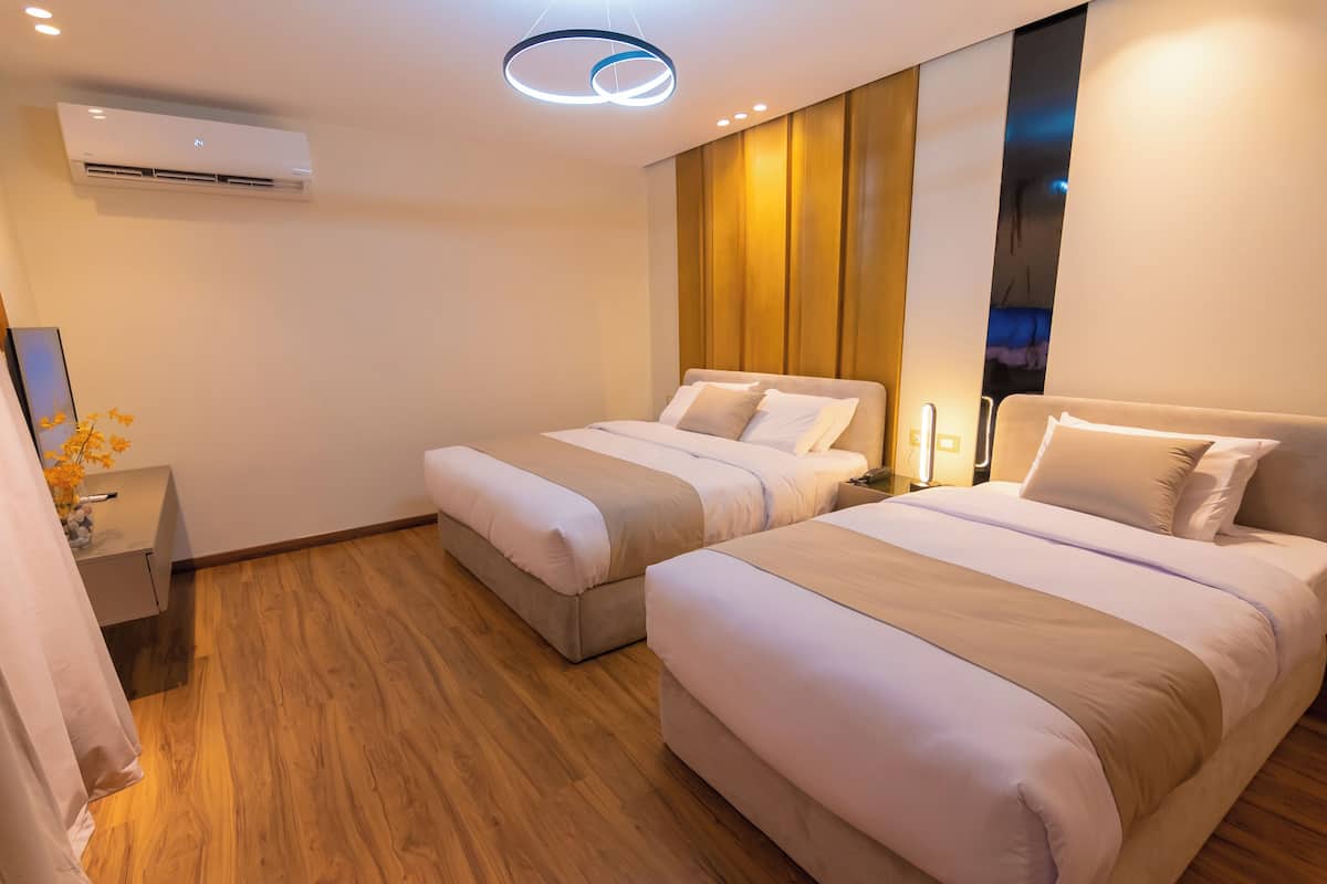 Moderni hotellihuone, jossa on kaksi sänkyä, puulattia, seinään asennettu ilmastointilaite ja kattovalaisin.