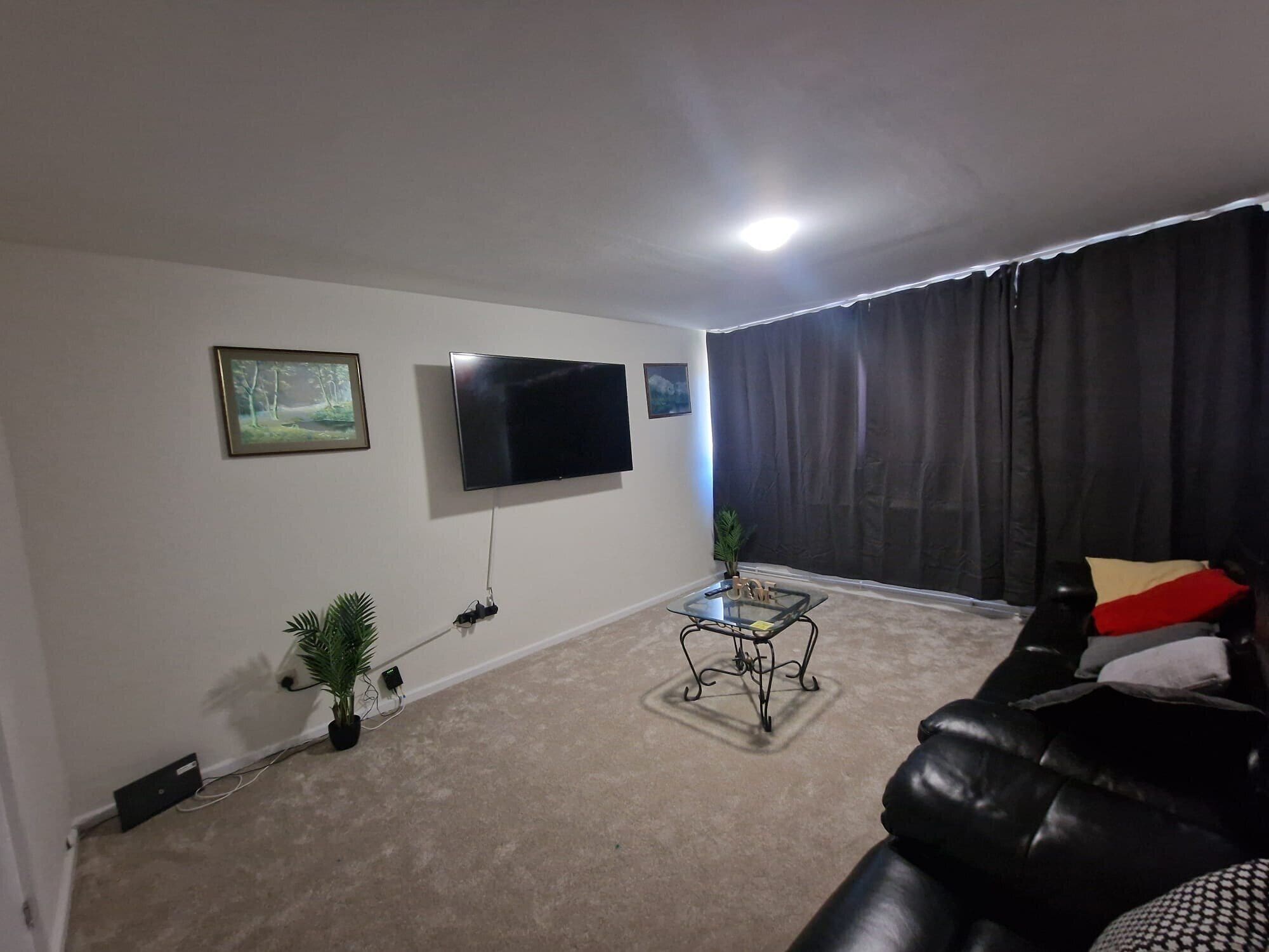 A living room with a flat-screen TV mounted on the wall, a black leather sofa, a glass coffee table, and a potted plant.