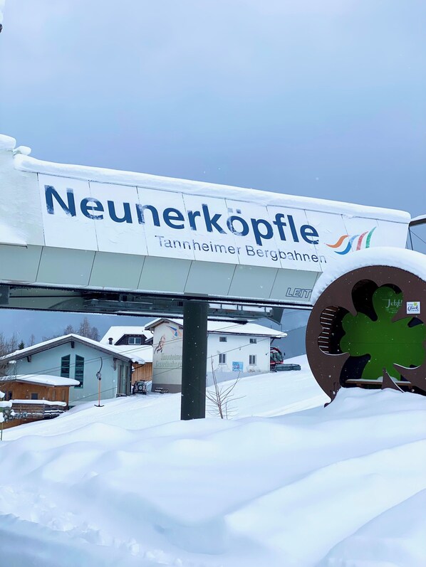 A sign for Neunerköpfle with a green shamrock logo, a snow-covered landscape, and a building with a snow-covered roof. - Lutz Alpenblick B&B (Schattwald)