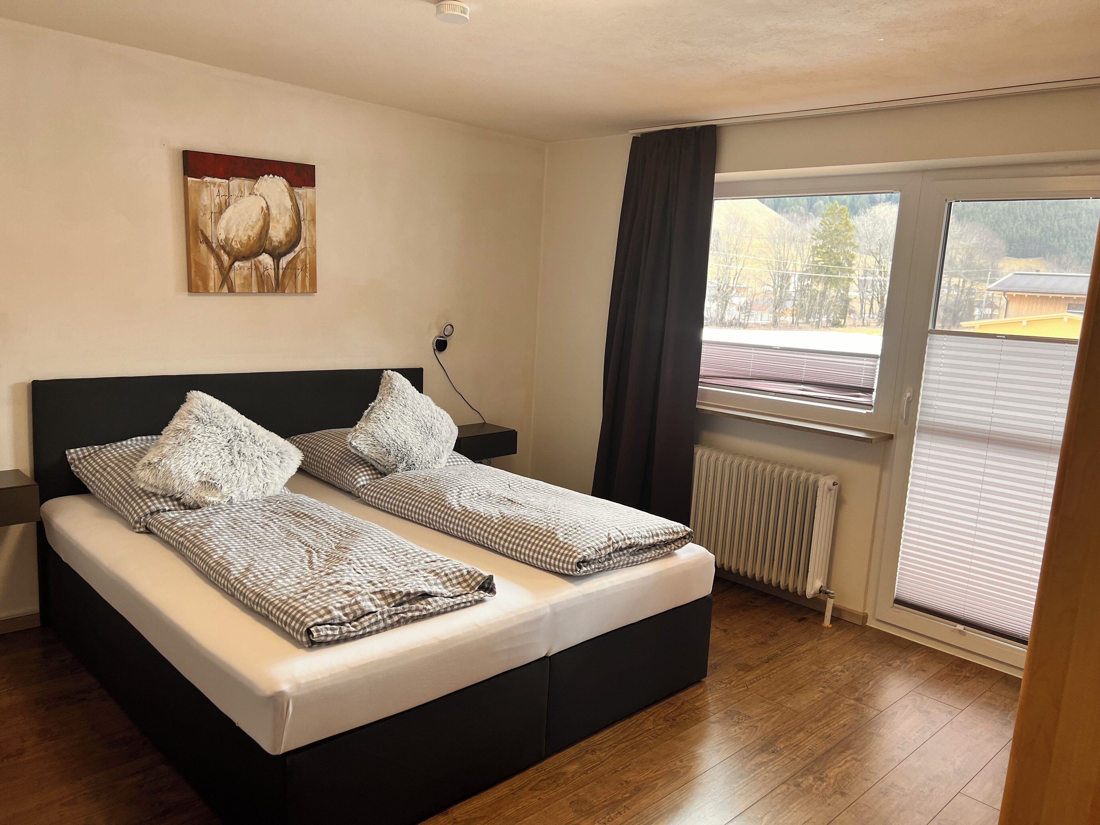 A bedroom with a bed, a nightstand, a painting, a window with curtains, and a radiator.