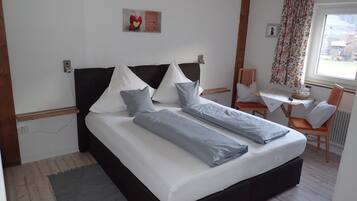 A neatly made bed with pillows and a blanket, a small table with a chair, and a window with curtains.