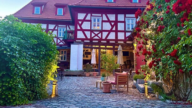 En traditionell byggnad med korsvirkesfasad och en innergård med kullersten, utomhusmöbler och krukväxter.