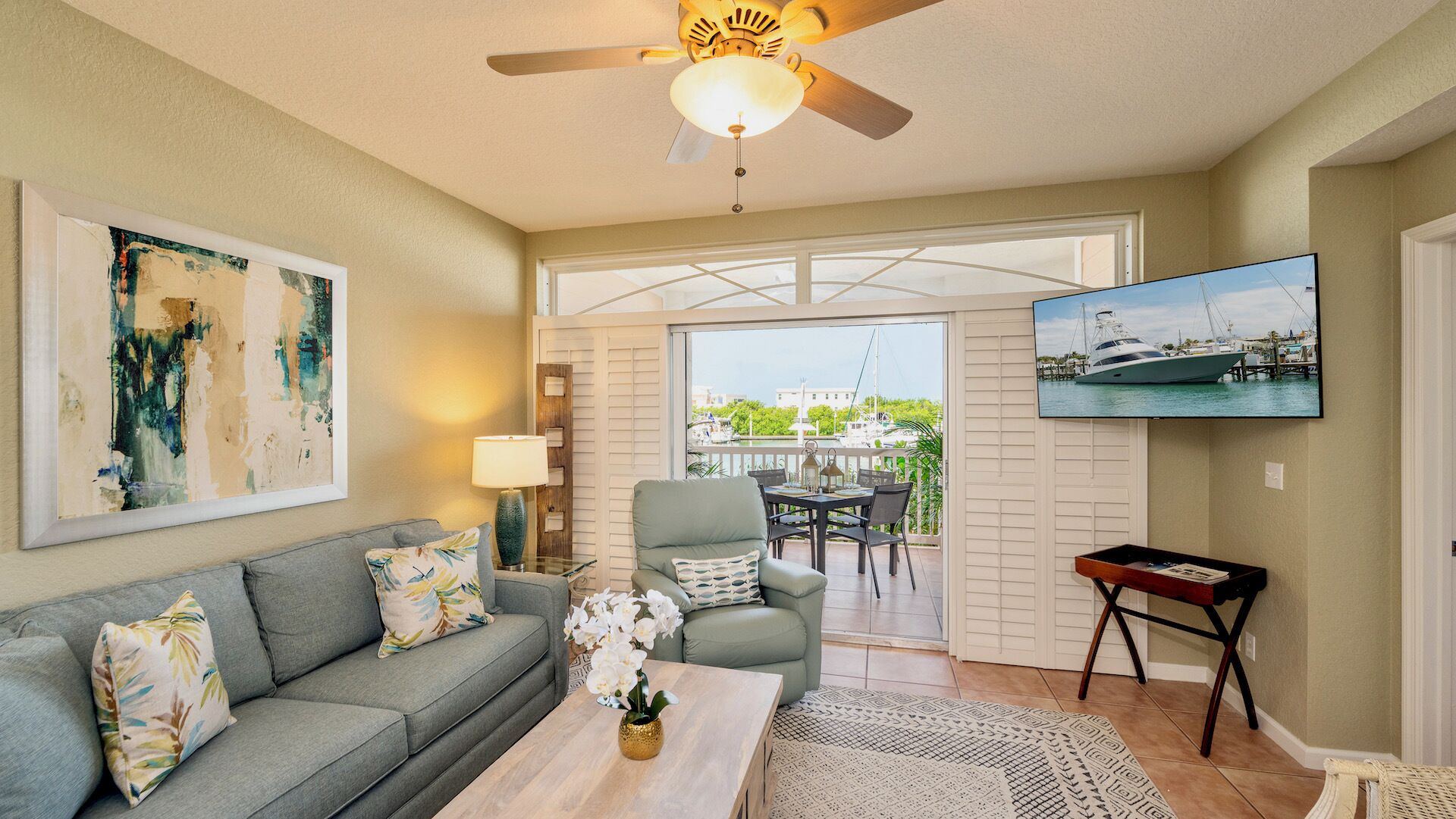 A living room with a gray sofa, a coffee table, and a TV on the wall. There is a ceiling fan, a window with a view of boats, and a door leading to an outdoor patio.