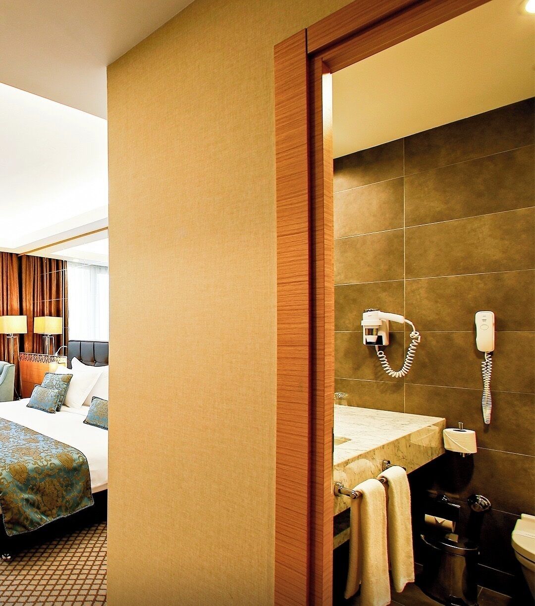 Una habitación de hotel con cama, lámparas de noche, baño con lavamanos, espejo y toallero.