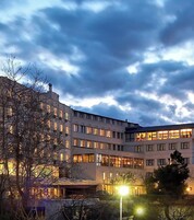 Un edificio de varios pisos con ventanas iluminadas, rodeado de árboles desnudos y un cielo nublado.