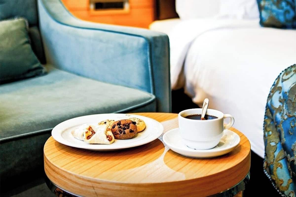 Ein Hotelzimmer mit einem Bett, einem Nachttisch mit Teller mit Essen und einer Kaffeetasse sowie einem blauen Sessel.