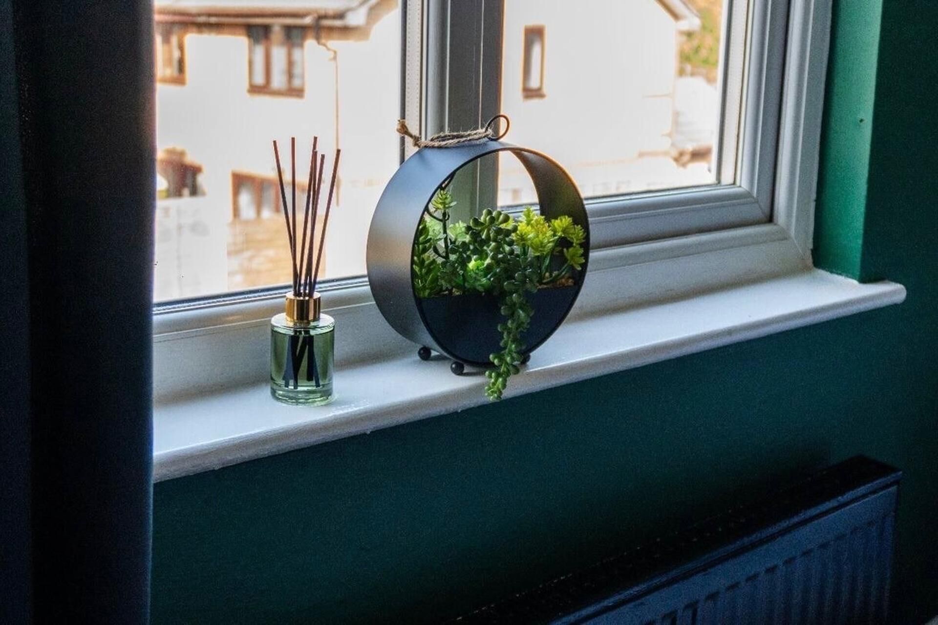 Un rebord de fenêtre sur lequel est posé un pot rond noir contenant des plantes vertes et un petit flacon en verre avec des tiges de roseau.