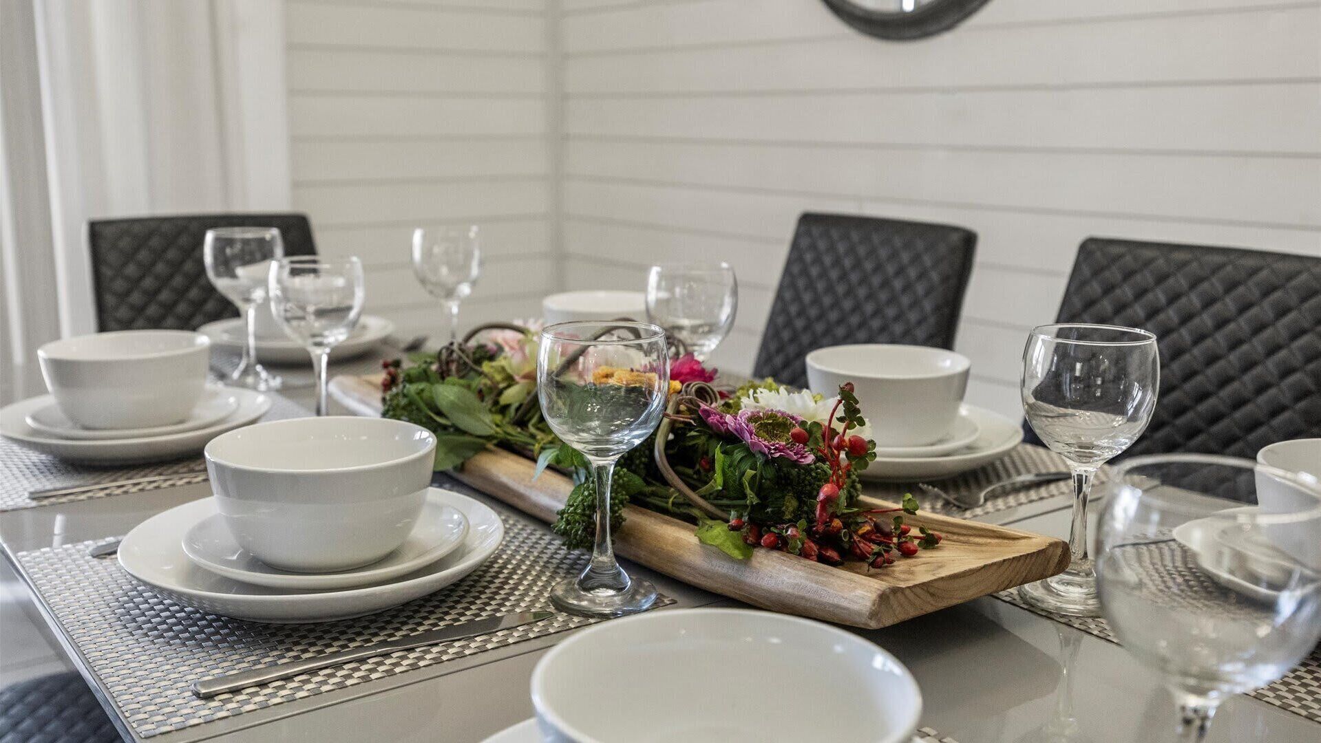 Een eettafel gedekt met witte kommen, borden en glazen, met een houten plank met bloemen en groen.