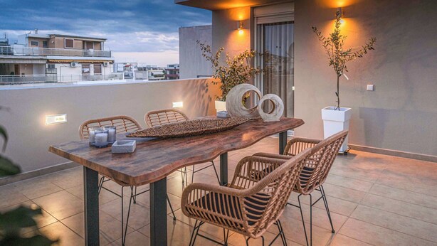 Een houten eettafel met rieten stoelen op een balkon, twee potplanten, en een uitzicht op gebouwen op de achtergrond.