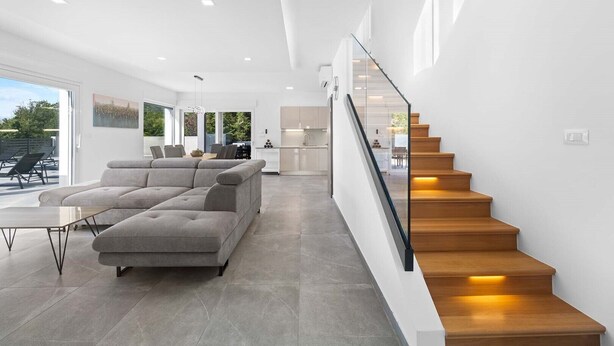 Un intérieur moderne avec un canapé d’angle gris, un escalier à marches en bois et, au fond, un espace cuisine.
