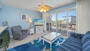 A living room with a blue sofa, a grey armchair, a glass coffee table, a ceiling fan, a TV, and a view of the ocean and a building named "Island".