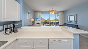 A modern kitchen with a central island, white cabinetry, a coffee maker, and a dishwasher. There is a dining area with a table and chairs, and a view of the outdoors through large windows.