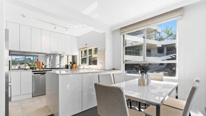 Une cuisine moderne avec des meubles blancs, un coin repas doté d’une table en verre et une vue sur un immeuble à travers de grandes fenêtres.