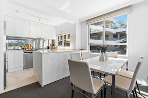 Una cocina moderna con armarios blancos, un comedor con mesa de vidrio y vistas a un edificio a través de amplios ventanales.