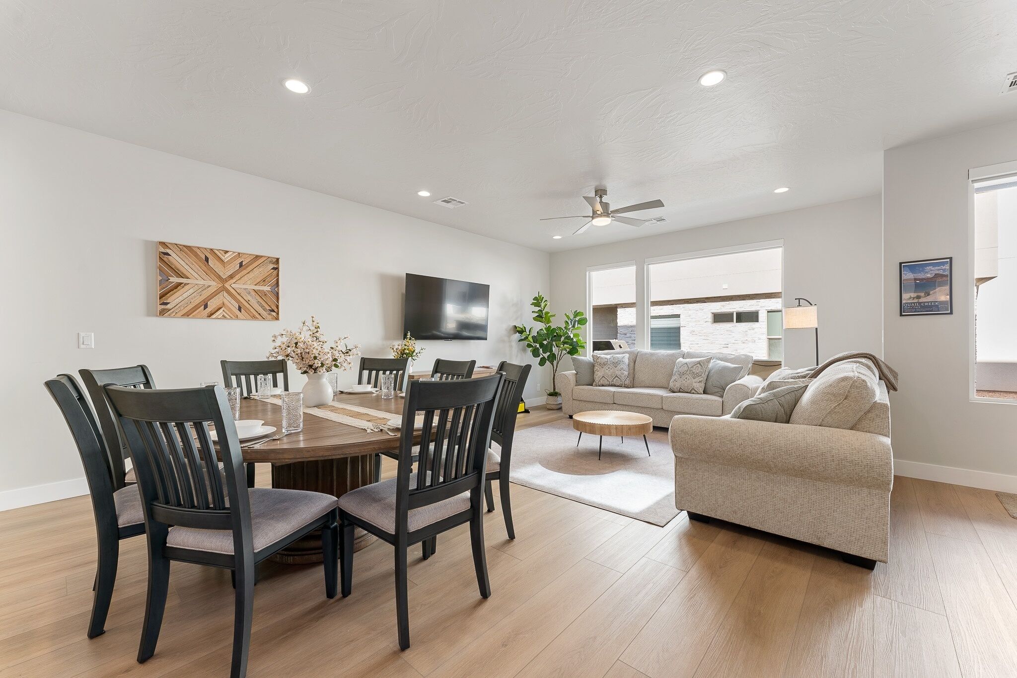 A modern living room with a dining area, a ceiling fan, and a large window providing natural light.