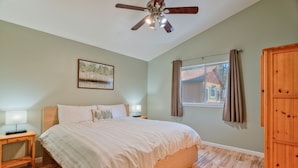 Une chambre à coucher avec un lit, un plancher en bois, un ventilateur de plafond, une table de chevet, une lampe, une fenêtre avec des rideaux et un tableau au mur.