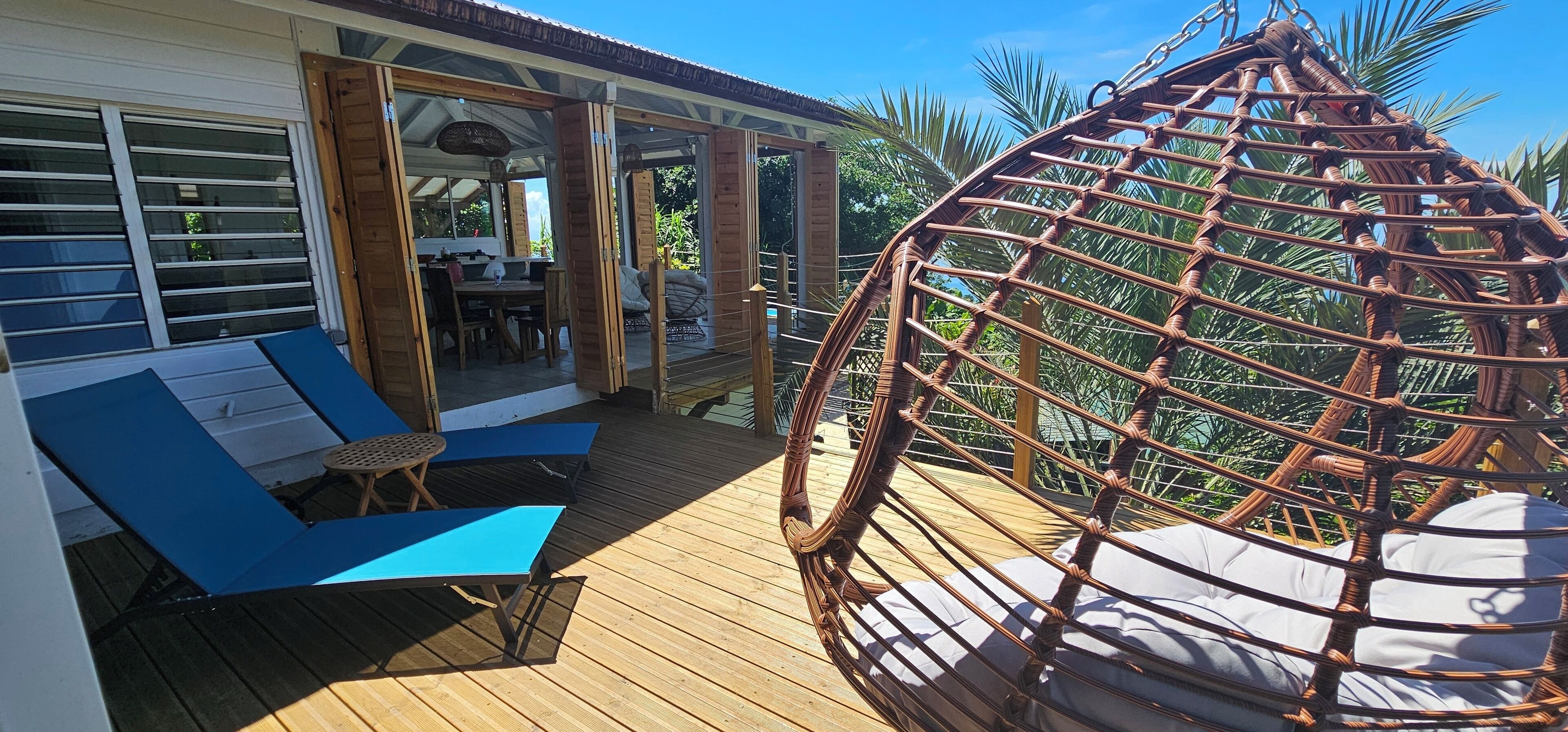 Une terrasse en bois avec des chaises longues bleues et un fauteuil suspendu en osier.