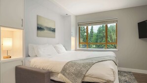 Quarto moderno com uma cama grande, uma mesa de cabeceira com abajur, uma janela com vista para árvores e uma TV na parede.