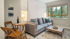Uma sala de estar moderna com um sofá cinzento, cadeiras de madeira, uma mesa de centro e uma janela grande com vista para árvores.